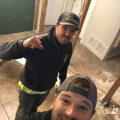 Two people wearing caps and dark hoodies smiling in an interior room undergoing renovation, with one pointing upwards.