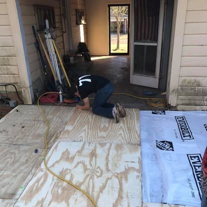A person kneeling on a wooden subfloor while installing plywood sheets on a porch.