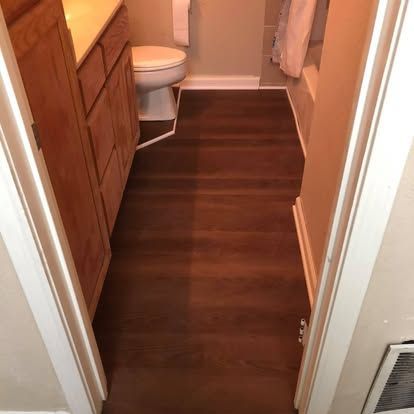 A small bathroom with dark wood-look vinyl flooring, a wooden vanity, a white toilet, and a bathtub with a white shower curtain.
