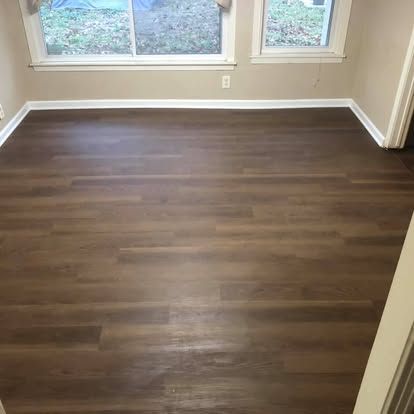 A room with dark brown wood-look flooring and white trim, featuring two windows overlooking a yard.