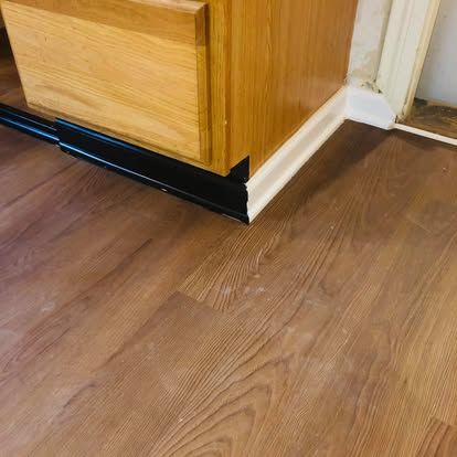 A wood-toned cabinet base meets a white baseboard, showing a sharp transition between black and white trim on a wood floor.