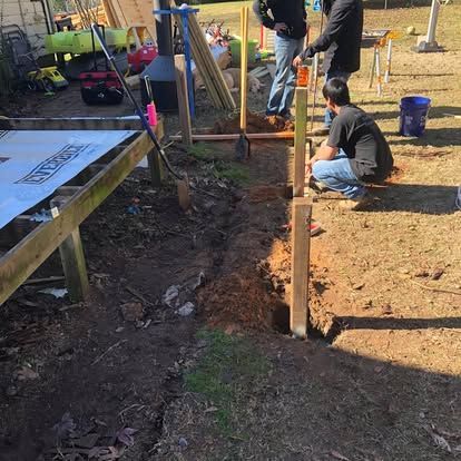 Two people work on a wooden deck project in a yard, one kneeling by a post hole in the dirt and one standing nearby.
