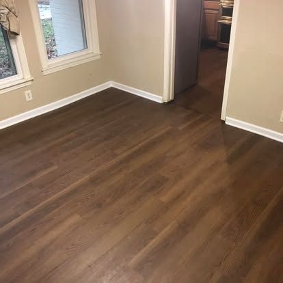 An interior view of a room with dark wood-look plank flooring, beige walls, white trim, and a doorway leading to a kitchen.