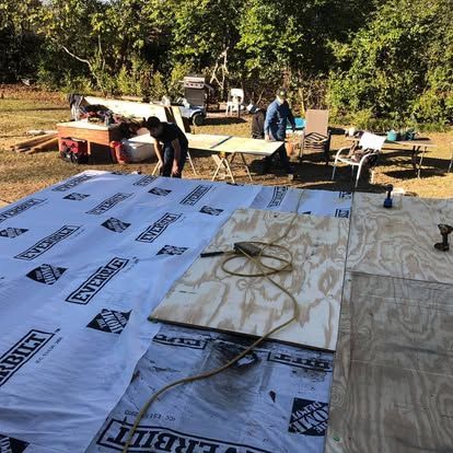 Two workers building a wooden deck outdoors, installing plywood subflooring over a white protective membrane.