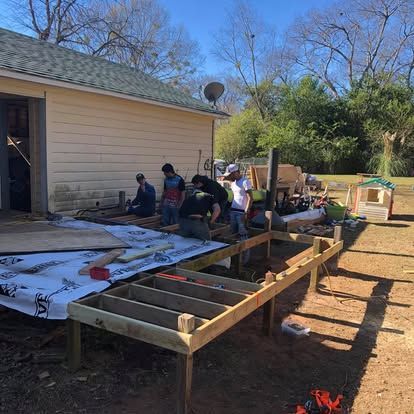 A group of people works together to build a wooden deck frame outside a house on a sunny day.