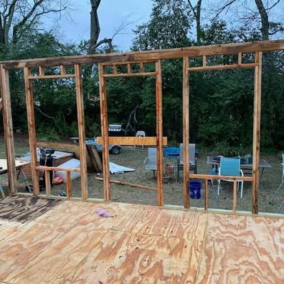 A wooden wall frame under construction, featuring three open sections for windows or doors, sits on a plywood floor outdoors.