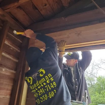 Two people in work clothes repair a wooden structure, with one person hammering a beam and the other assisting nearby.