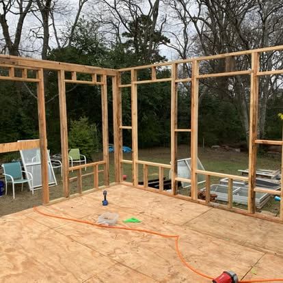 Wooden wall framing for a new structure built on a plywood subfloor, set against a background of trees.