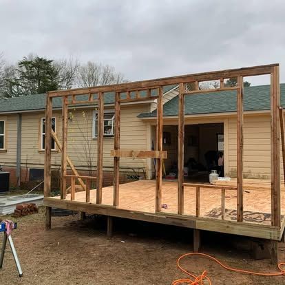 The wooden frame of a new building addition stands on a plywood floor in front of an existing yellow house.