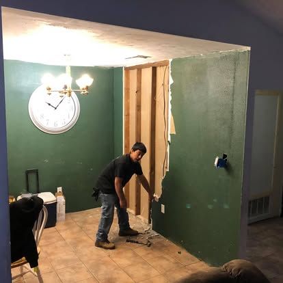 A person works in a room with green walls, removing a section of drywall to expose the wooden wall studs.