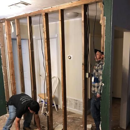 Two people work on a wall renovation, with one kneeling by exposed studs and another standing beside hanging wires.