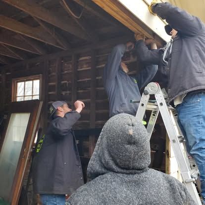 Four people work together on a ladder and scaffolding to repair the exterior frame of a weathered wooden building.