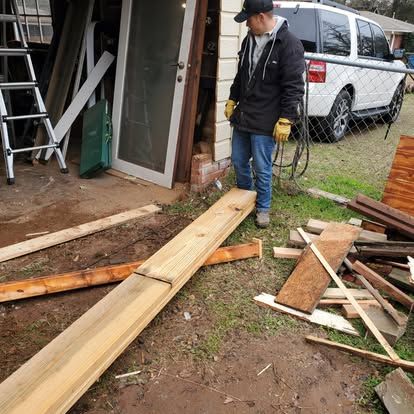A man in a black jacket and yellow gloves stands next to a long wooden board in an outdoor workspace near a white SUV.