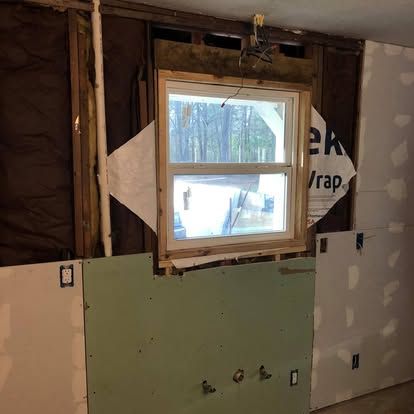 A partially renovated wall with a centered window, exposed studs, insulation, white house wrap, and installed drywall.