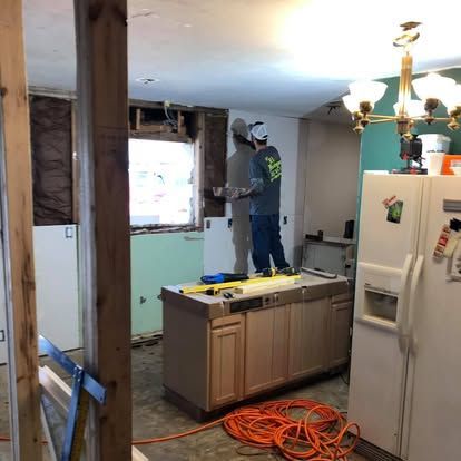 A person in a gray shirt and hat installing drywall in a kitchen renovation, with a refrigerator and kitchen island nearby.