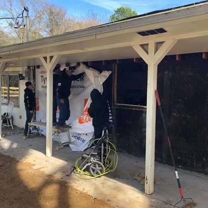Three people work on a house exterior, attaching a Tyvek house wrap barrier to the studs under a covered porch.