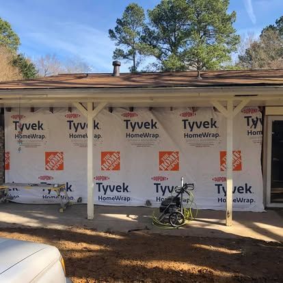 Exterior wall of a house under renovation, covered in white Tyvek HomeWrap, with a porch, roof, and trees in the background.