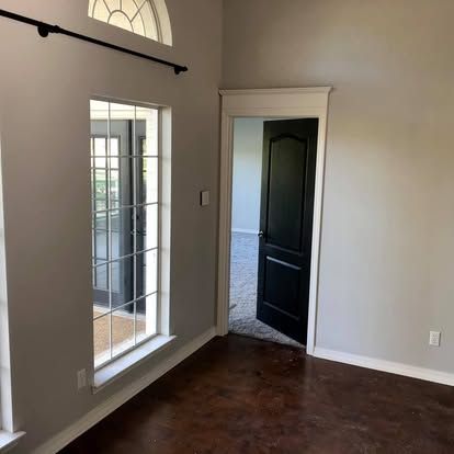 A room with grey walls, a large window, a black door leading to another room, and dark-stained concrete floors.