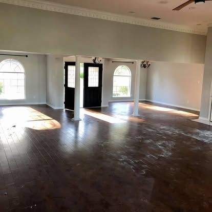 A spacious, empty room with dark wood flooring, high ceilings, a black double door, and arched windows letting in light.