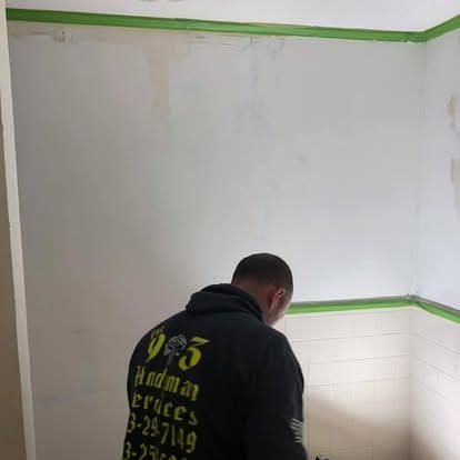 A person in a branded black hoodie performs repair work on a room wall with green painter's tape along the ceiling and tile.