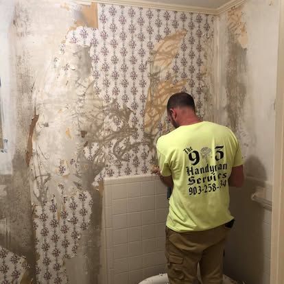 A worker in a neon yellow shirt removes old, patterned wallpaper from a bathroom wall above a tiled backsplash.