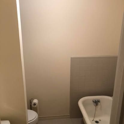 A small bathroom showing a white toilet, a roll of toilet paper, and the end of a white claw-foot tub against a tiled wall.