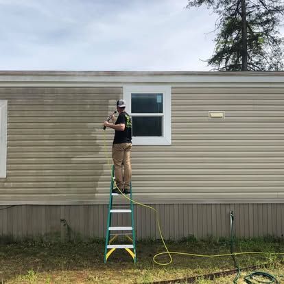 A person standing on a green ladder power washes dirty tan siding on the side of a house, leaving a clean streak.