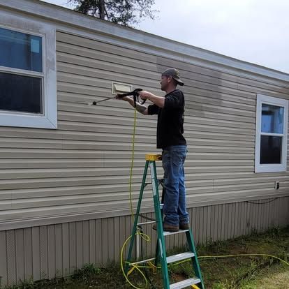 A person on a stepladder uses a pressure washer to clean the light-colored vinyl siding of a mobile home.