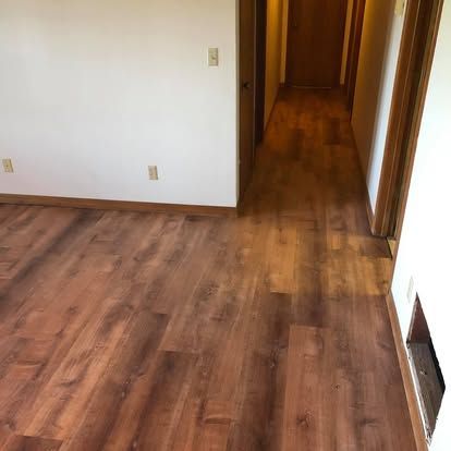 An empty room with dark wood flooring, white walls, wood trim, and a hallway leading to a door in the background.