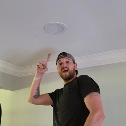 A person in a black t-shirt and baseball cap smiles while pointing up at a recessed light fixture in a white ceiling.