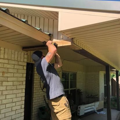 A person wearing a grey shirt and cap uses a power drill on the wooden eave of a house with light-colored brick walls.