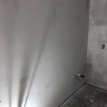 Interior room under construction with gray drywall, exposed plumbing pipes on the wall, and an electrical outlet.