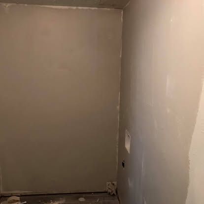 A small, enclosed room with gray, unfinished drywall panels and an electrical outlet on the right wall.