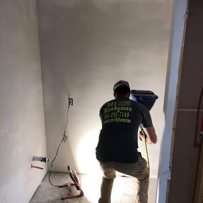 A contractor in a branded t-shirt uses a drywall hopper gun to apply texture to a wall in a room under construction.