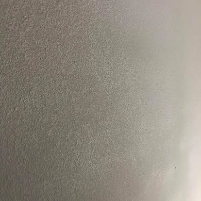 A close-up, flat texture of a wall painted in a light, neutral gray or beige with a slightly gritty, matte finish.
