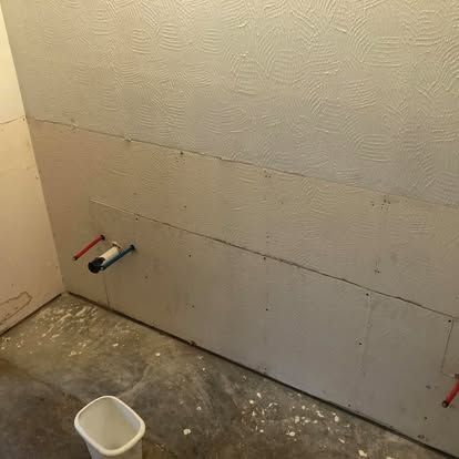 A wall under renovation with exposed drywall, plumbing pipes extending from the center, and a white bucket on the floor.
