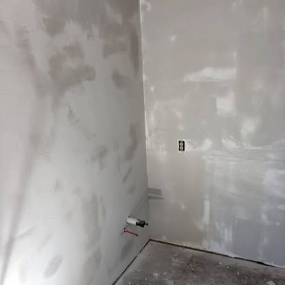 Two drywall-finished walls meet at a corner, featuring a partially installed electrical outlet and protruding pipes.