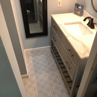 A bathroom vanity with a white marble countertop and gray drawers, featuring patterned floor tiles and a wall mirror.