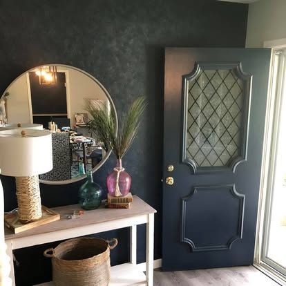 An entryway featuring a dark navy door, a round mirror, and a console table with a textured lamp and woven basket.