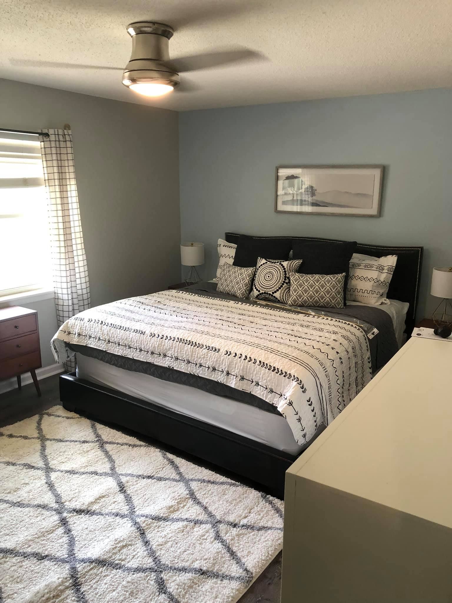 A bedroom with a bed featuring dark headboards, a patterned throw, a large rug, a ceiling fan, and soft blue walls.