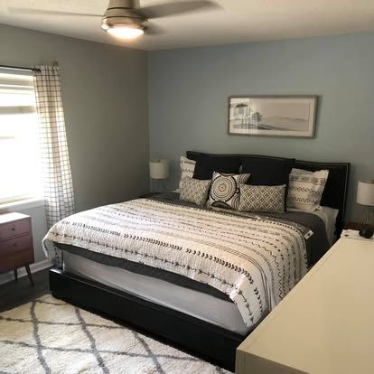A bedroom with a bed featuring a patterned white and black comforter, a black headboard, blue-grey walls, and a rug.