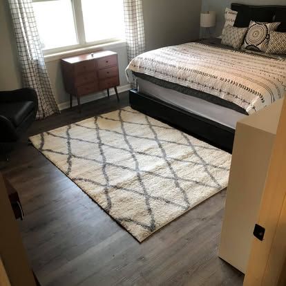 A bedroom with wood flooring, a bed featuring a patterned quilt, a cream-colored trellis rug, and a small wooden dresser.