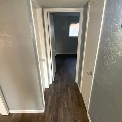 A hallway with light gray walls, dark wood-look flooring, and white trim leads to an open doorway showing a distant room.