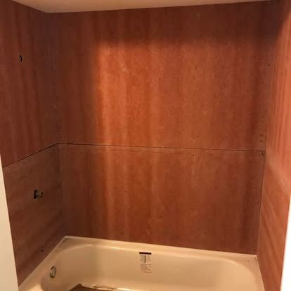 An unfinished shower alcove with brown plywood walls installed around a white bathtub.