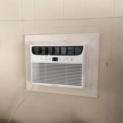 A white window air conditioner unit installed in a wall with a white rectangular border on a paneled wall.
