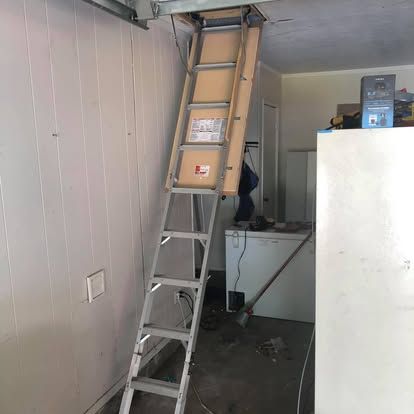 A metal pull-down attic ladder extended from a ceiling hatch into a garage, positioned next to a white chest freezer.