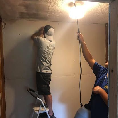 Two people work on a wall, with one person on a ladder and another holding a spray device towards the ceiling.