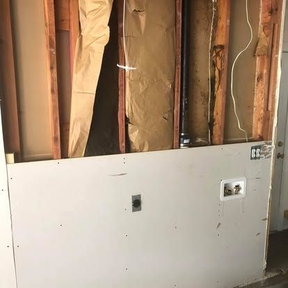 A wall under renovation with lower drywall installed, revealing wooden studs, insulation, and plumbing fixtures.