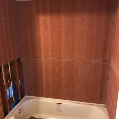 A partially renovated bathroom alcove featuring plywood-covered walls above a white bathtub with exposed plumbing.