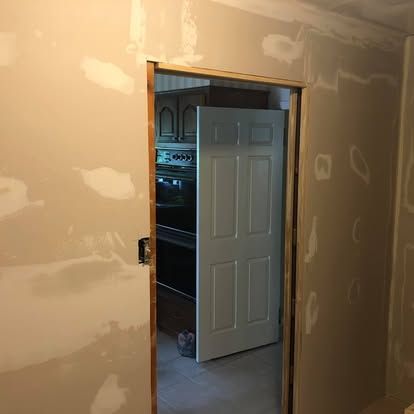 An unfinished wall with drywall mud patches around a doorway looking into a kitchen with a white door and oven.